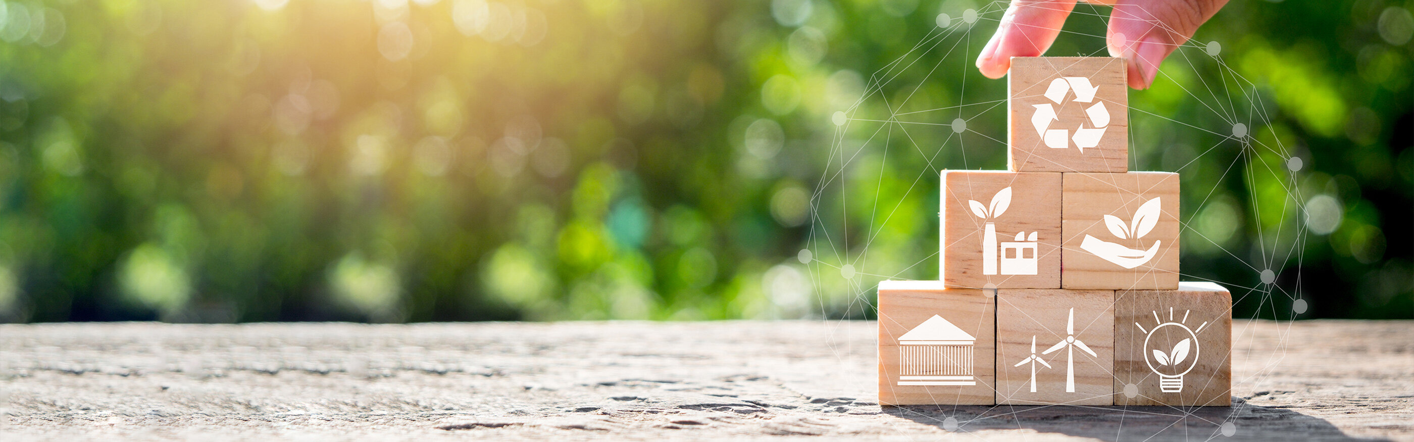 Wooden blocks with sustainability symbols are piled up to form a pyramid, green blurry nature background